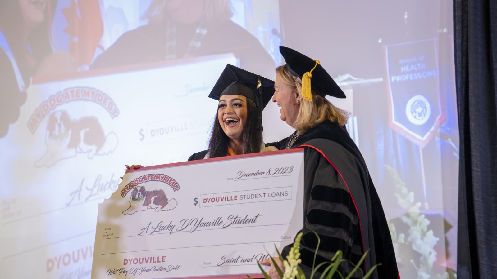 President Dr. Lorrie Clemo, in graduation regalia, stands with student in graduation cap and gown. They hold a giant check representing a payment of the lottery winner's student loan debt.
